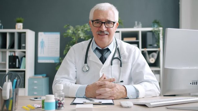 Smiling doctor wearing white gown and glasses looking at camera sitting at desk in hospital office room. Modern medicine and senior people concept.