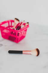 beauty industry products concept, pink shopping basket with variety of make-up brushes in it for face and eye make-up and one brush outside in the foreground