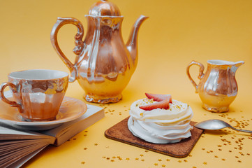 Bright and beautiful snack. White cake made of meringue, cream and fresh strawberries and Golden tea utensils: a mug, a teapot and a container for cream on a bright yellow background with gold stars