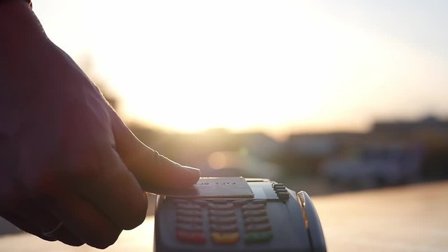 A men holds a credit card in her hand in front of the terminal in a cafe near the window against the sunset. Credit card without image. Credit card payment at the terminal