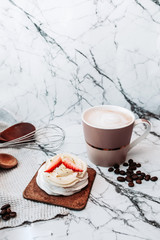 White dessert of meringue, cream and fresh strawberries, hot drink mug, open book and coffee beans on white marble surface. Vertical image