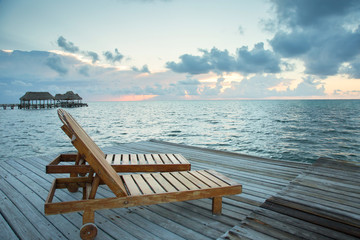 Wooden lounge chairs at the end of the dock with fluffy clouds and pink setting sun. Calm ocean in background