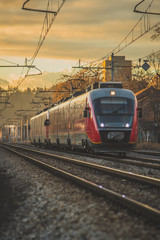 European style red passenger train of a modern type on a straight track in an urban setting during evening hours. Romantic railway journey at a sunset. Romantic evening commute. © Anze