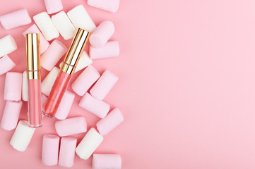 Obraz na płótnie Canvas Pink pastel lip gloss on a background of sweets. Decorative women's cosmetics accessory on a light backdrop with marshmallows. Makeup for lips with sparkles in transparent packaging with copy space