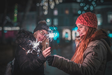 Two young women with sparklers in their hands enjoying an evening in the christmas lights lit city centre. Cold winter atmosphere and enjoyment outside in urban area.