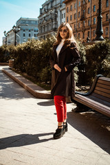 Pretty business young woman model in fashionable sunglasses in a trendy coat stands on road in the city. Sexy girl posing outdoors. Modern lady.