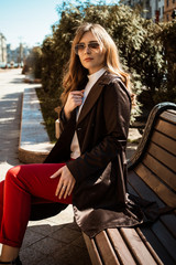 Outdoor fashion details. Young brunette woman in coat sitting on a bench in the city. Sunny spring day.