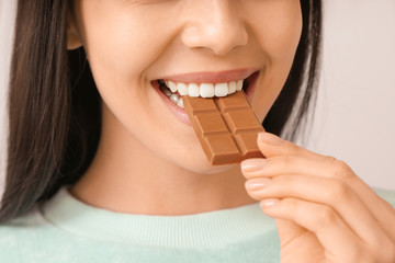 Beautiful young woman eating tasty chocolate on light background, closeup