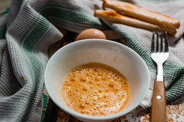 Whipped with a fork and mixed raw egg broken into a white bowl with yolk and protein, next to the shell, fork and towel on a rusty metal background as at home in the village, everything simple and ord