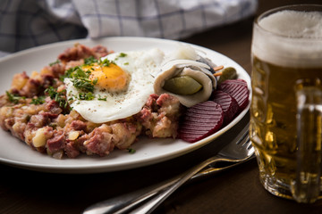 Norddeutscher Hamburger Labskaus mit Rindfleisch Corned Beef, Rote Beete, Kartoffeln, Saure Gurken, Spiegelei, Rollmops und Bier auf Holztisch
