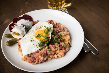 Norddeutscher Hamburger Labskaus mit Rindfleisch Corned Beef, Rote Beete, Kartoffeln, Saure Gurken, Spiegelei, Rollmops und Bier auf Holztisch