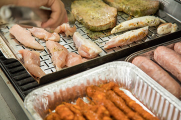 Mann wendet und grillt Hühnchen Filet, Schweine Nackensteak, frische rohe Bratwurst und Bacon Speck Pflaumen auf Gasgrill in Edelstahl Grillkorb Pfannen