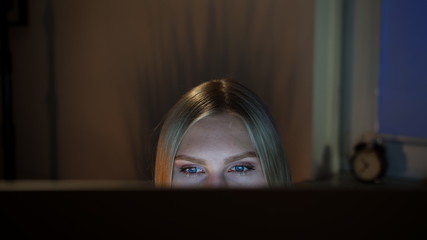 Crop view of beautiful female sitting at night at computer and looking at camera over top of...