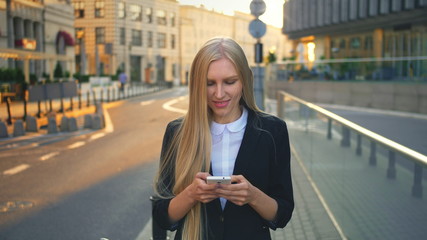 Obraz premium Elegant blond woman in suit walking on street and browsing smartphone with smile against urban background.