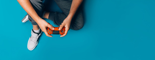 A young guy plays video games in his hands holding a red gamepad on a blue background, sitting in gray sneakers, long banner - Powered by Adobe