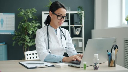 Portrait of young female doctor in white uniform working with laptop computer sitting at desk in office. Modern technology and medicine concept. - Powered by Adobe