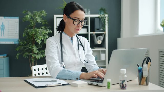 Portrait of young female doctor in white uniform working with laptop computer sitting at desk in office. Modern technology and medicine concept.