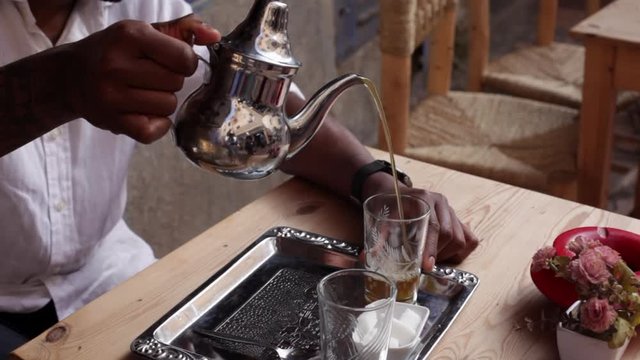 Male hand pouring tea from ornate silver teapot into glasses on small table according to Moroccan tradition. Ritual preparation of mint tea in Morocco