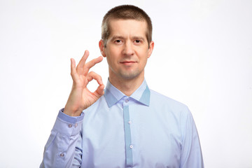 successful happy man shows the gesture OK with one hand, in a blue light casual shirt staring at the camera. Isolated