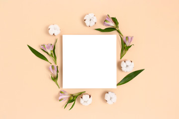 Square paper card mockup with frame made of green leaves, cotton, alstroemeria flowers on a beige background. Spring concept.