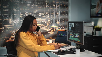 Mixed race colorist talking by smartphone and writing notes in modern production studio. In the backgrounds - skyscrapers. - obrazy, fototapety, plakaty
