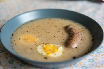 Sour soup or white borscht with white sausage and egg. Easter food. Traditional cuisine, in the background a tablecloth with a floral motif.