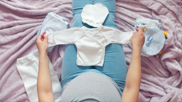A pregnant girl in jeans and a white t-shirt sits on the bed and puts out small baby things for the newborn. Pregnant mom prepares children's undershirt
