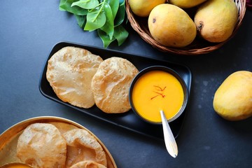 Indian Food - Aamras is a sweet dish made from the pulp of the Alphonso mango fruit. Served with hot puri/poori. Sweet Mango puree garnished with Kashmiri Saffron. with whole mangoes in background.