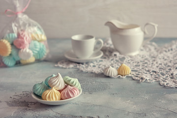 Multi-colored meringues in pastel shades with cups and a jug on a light textured wooden background. The concept of Breakfast with tea or coffee, home comfort and tenderness. Close-up, selective focus