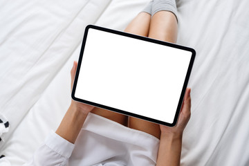 Top view mockup image of a woman holding black tablet pc with blank desktop white screen while lying down on a cozy white bed at home