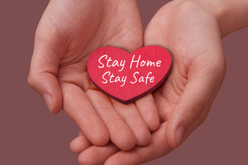 Women's hands hold a red wooden decorative heart with the inscription Stay Home Stay Safe. The concept of voluntary self-isolation, protection from the pandemic, slogan.