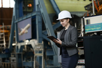 Portrait of a female factory manager in a white hard hat and business suit. Controlling the work process at the airplane manufacturer.
