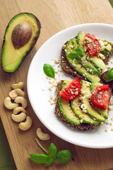 Avocado sandwich on a white plate with full grain bread, tomatoes, basil and cashew nuts. Wooden background.