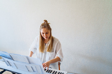 Woman is quarantined at home coronavirus. Attractive girl in a white shirt and jeans staying at home on self-isolation and playing the electronic piano - synthesizer. Hobbies - what to do at home?