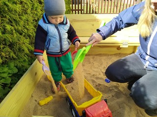 A little boy with mom pouring sand into toy truck in the sandbox surrounded by greenery on a sunny day