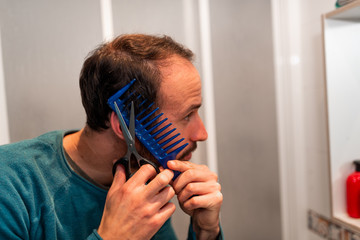 young man cutting his own haircut with scissors during quarantine at home, taking care of his look