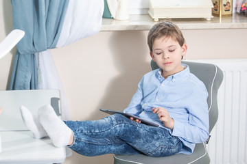 lovely 7yo boy in blue shirt sits at the desk with his legs on the table looking at the tablet working from home
