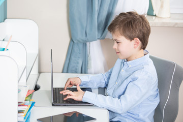 work from home - smiling young boy in blue shirt sits at his desk working on his notebook computer