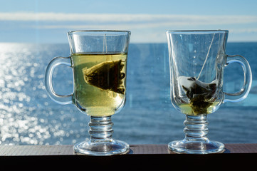 Two transparent glass cups with a bag of green leaf tea on a background of the sea.