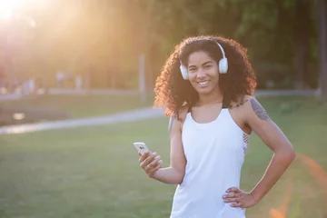 Fotobehang Sympathisch lachende junge Frau in der Abendsonne, sie trägt Sportkleidung und Kopfhörer und hat ein Smartphone in der Hand  © Wellnhofer Designs