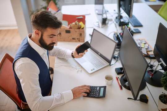 young business man using calculator at his desk in office, processing data, holding cell phone in front of computer. business , analyzing, processing data, calculating