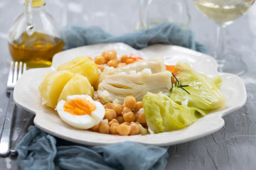 boiled cod fish with boiled vegetables and egg on white dish and glass of wine on ceramic background