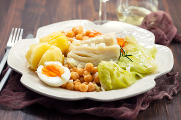 boiled cod fish with boiled vegetables and egg on white dish and glass of wine on ceramic background