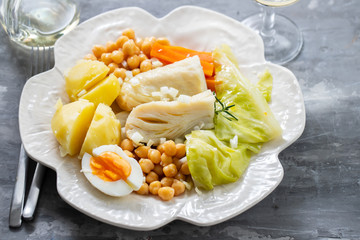 boiled cod fish with boiled vegetables and egg on white dish and glass of wine on ceramic background