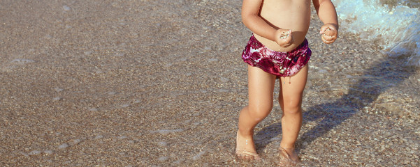 little girl in purple swimming trunks 
on the beach carries sea pebbles and seashells in her hands. Rest and travel. Vacation concept with little children. People and advertising concept. Childhood. 