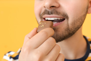 Handsome young man eating tasty chocolate on color background, closeup