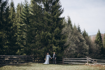 Stylish wedding couple in the mountains. A guy in leather jacket and young girl in gray blue wedding dress, denim jacket and hat stand on the background of forest, mountains and a wooden fence