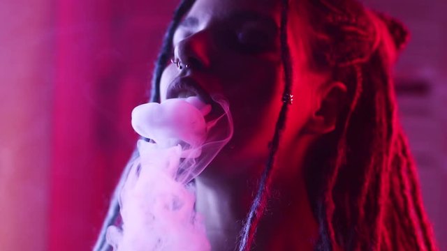 exhales smoke closeup girl with dreadlocks. red and blue color. vape.