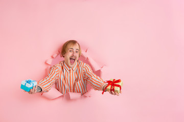 Giving gifts, presents. Cheerful caucasian young man poses in torn coral paper background, emotional expressive. Breaking on, breakthrought. Concept of human emotions, facial expression, sales, ad.