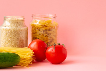 Noodles, rice, pasta in glass jars stand on a pink background. Nearby are red tomatoes and green cucumber. Raw materials for cooking.